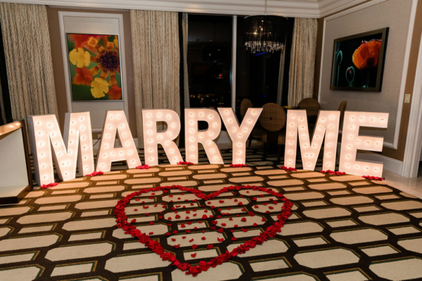 A Las Vegas hotel room is decorated for a marriage proposal. A heart formed from rose petals on the carpet, and large light-up marquee letters spell "MARRY ME".