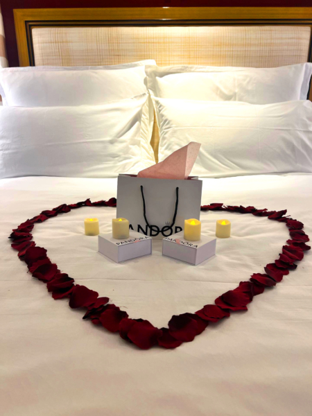 A bed in a hotel room is decorated with a rose petal heart and gifts in the center. The decor looks romantic and could be for an anniversary or marriage proposal