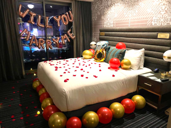A Las Vegas hotel room is decorated for a marriage proposal. Balloons, rose petals and a "will you marry me" banner decorate the bed, floor and window. The Las Vegas strip is seen outside through the window.