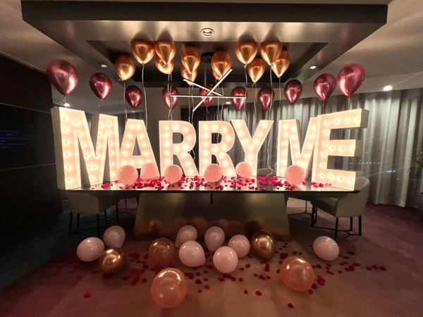 A Las Vegas hotel room with romantic decor for a marriage proposal. Balloons and "MARRY ME" light up marquee letters are set on the table and floor