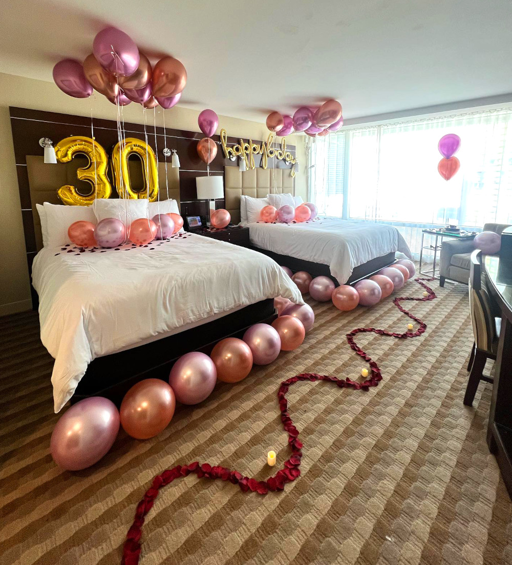 A room at a hotel in Las Vegas is decorated for a romantic birthday celebration. Balloons are set up on and around the bed, with rose petals decorating the floor.