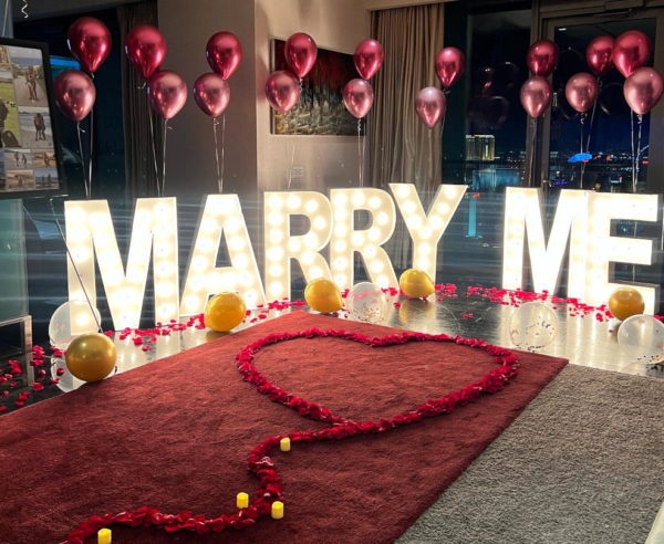 A Las Vegas hotel room with romantic decor for a marriage proposal. Balloons, rose petals and "MARRY ME" light up marquee letters are set up